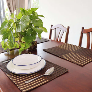 Dining table set with plates, spoons, and placemats on a wooden table with a plant in the background.