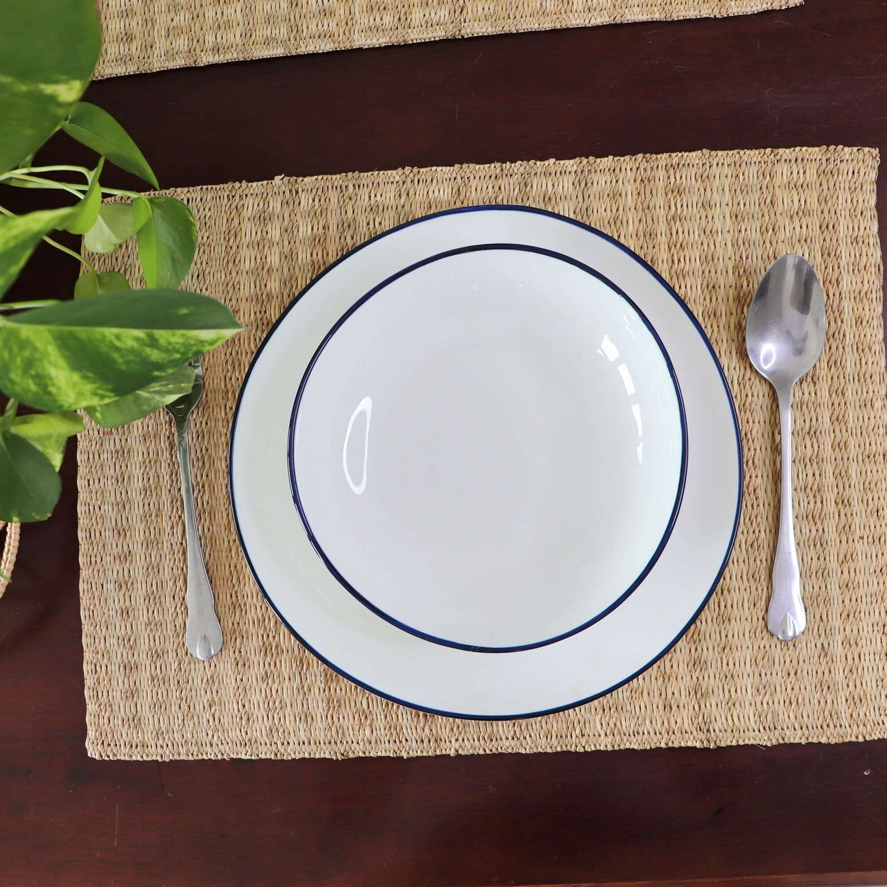 White plate with blue rim on a woven placemat, accompanied by a spoon and fork, on a wooden surface.