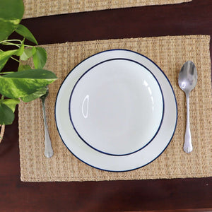 White plate with blue rim on a woven placemat, accompanied by a spoon and fork, on a wooden surface.