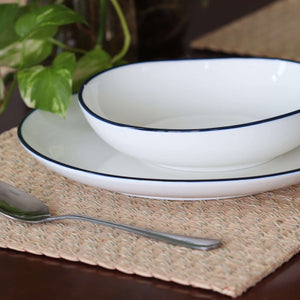 White ceramic plates with blue rims on a woven placemat, with a plant in the background.