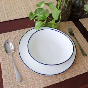 White bowl with blue rim on a woven placemat, accompanied by a spoon and fork.