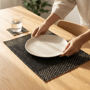 Person placing a plate on a Lavila Handwoven Placemat on a wooden table, showcasing elegant dining decor.